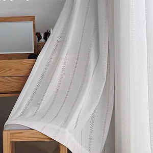 May include: White sheer curtains with vertical patterned stripes. The curtains are hanging in front of a wooden vanity with a mirror. The sheer fabric allows light to pass through, creating a bright and airy atmosphere.