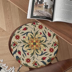 May include: A round chair cushion with a cream background and a colorful floral pattern. The pattern features red, yellow, blue, and green colors. The cushion is made of fabric and has a circular shape.
