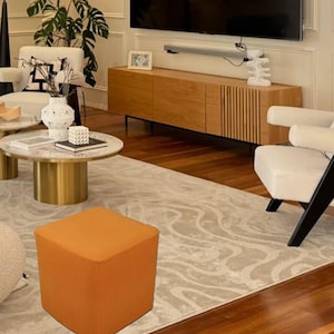 Puede incluir: Un puf cuadrado naranja con una cubierta de tela texturizada. El puf está sentado en una alfombra de color crema en un ambiente de sala de estar.