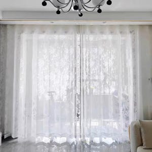 Puede incluir: Cortinas blancas transparentes con un delicado estampado floral. Las cortinas cuelgan frente a una ventana, permitiendo el paso de la luz. El diseño incluye un borde decorativo. Un candelabro es visible en la parte superior de la imagen.