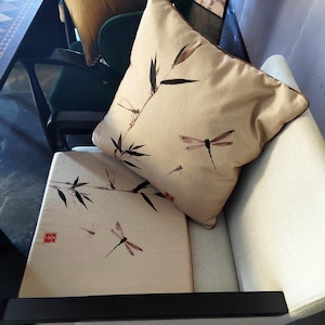 Può includere: Una sedia beige con una struttura nera e un cuscino beige con un motivo a libellula e bambù. Un secondo cuscino in tessuto dorato con bordatura marrone è sulla sedia.