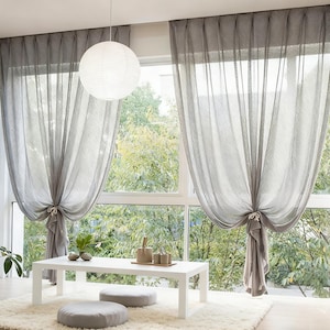 May include: Sheer gray curtains draped on either side of a large window, tied back with fabric. A white paper lantern hangs from the ceiling. A white table and two gray floor cushions sit on a white rug.