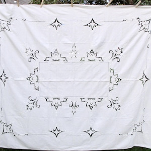 May include: A white rectangular tablecloth with intricate black floral and geometric cut-out designs. The tablecloth is hanging outside on a clothesline, showcasing its full size and details. The design includes a central rectangular pattern and corner accents.