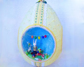 May include: A yellow Easter egg with a white lace design and a blue and white interior scene. The scene includes a white bunny holding a drum, a green and yellow bird, and a string of colorful lights.