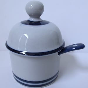 May include: A small, white ceramic sugar bowl with a lid and a handle. The bowl and lid are accented with dark blue stripes and trim. The lid has a small, round knob. The handle is also dark blue.