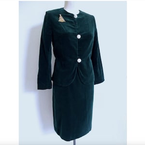 May include: A dark green velvet skirt suit displayed on a mannequin. The jacket has a round collar, three large, round, white buttons, and a gold-colored Christmas tree brooch. The skirt is a straight, knee-length style.