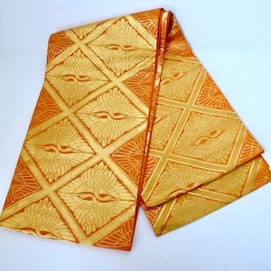 May include: An orange and gold textile featuring a repeating pattern of stylized birds within diamond shapes. The fabric is folded, showcasing the intricate design and texture. The textile has a luxurious appearance.