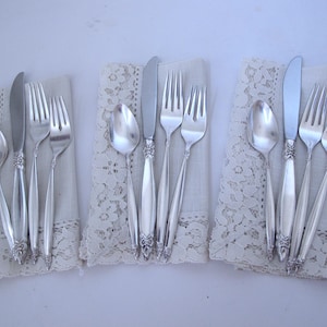May include: Three place settings of silver cutlery, each with a spoon, knife, and two forks, presented on decorative lace napkins. The cutlery features ornate handle detailing. The setting is on a white surface.