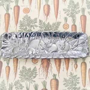 May include: A rectangular, silver-colored metal tray with a detailed relief of several rabbits nestled among leaves. The background features a carrot-themed pattern with botanical illustrations and text.