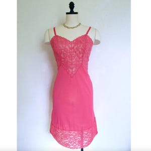 May include: A vintage pink slip dress with delicate lace detailing. The bodice features a lace overlay, and the hemline is trimmed with matching lace. The dress has thin spaghetti straps and is displayed on a mannequin.