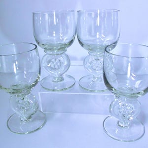May include: Four clear glass goblets. Each goblet has a rounded bowl, a decorative stem with a heart-shaped design, and a circular base. The goblets are arranged on a white surface, with a light blue background.