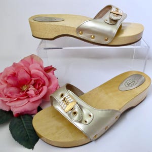 Könnte beinhalten: Zwei Paar goldfarbene Clogs mit Holzsohlen. Die Clogs haben einen metallischen Riemen mit dekorativer Schnalle. Die Holzsohlen sind hellbraun. Die Clogs werden auf einem transparenten Ständer mit einer rosa Rose und grünen Blättern präsentiert.