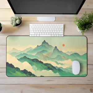 May include: A rectangular mouse mat with a landscape painting of layered green and blue mountains and a red sun. A white computer mouse and keyboard are on the wooden desk.