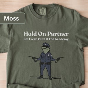 May include: Moss green t-shirt with the text "Hold On Partner I'm Fresh Out Of The Academy." A cartoon frog in a police uniform is holding two guns. The shirt is made of a soft material.
