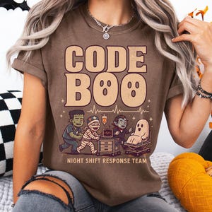 May include: A brown t-shirt featuring the text CODE BOO and a graphic of a Frankenstein monster, a mummy, a vampire, and a ghost around a medical cart. Below the characters, the text reads NIGHT SHIFT RESPONSE TEAM with a heartbeat line.