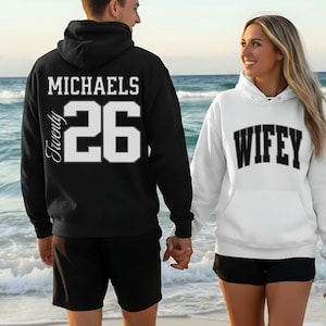 May include: A black hoodie with the name "MICHAELS" and the number "26" in white. A white hoodie with the word "WIFEY" in black. The couple is holding hands on a beach.