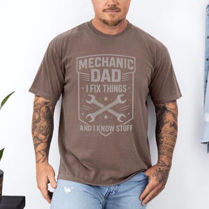 May include: A brown t-shirt with a gray graphic that reads "MECHANIC DAD - I FIX THINGS - AND I KNOW STUFF." The graphic includes crossed wrenches and stars. The shirt has short sleeves and a crew neck.
