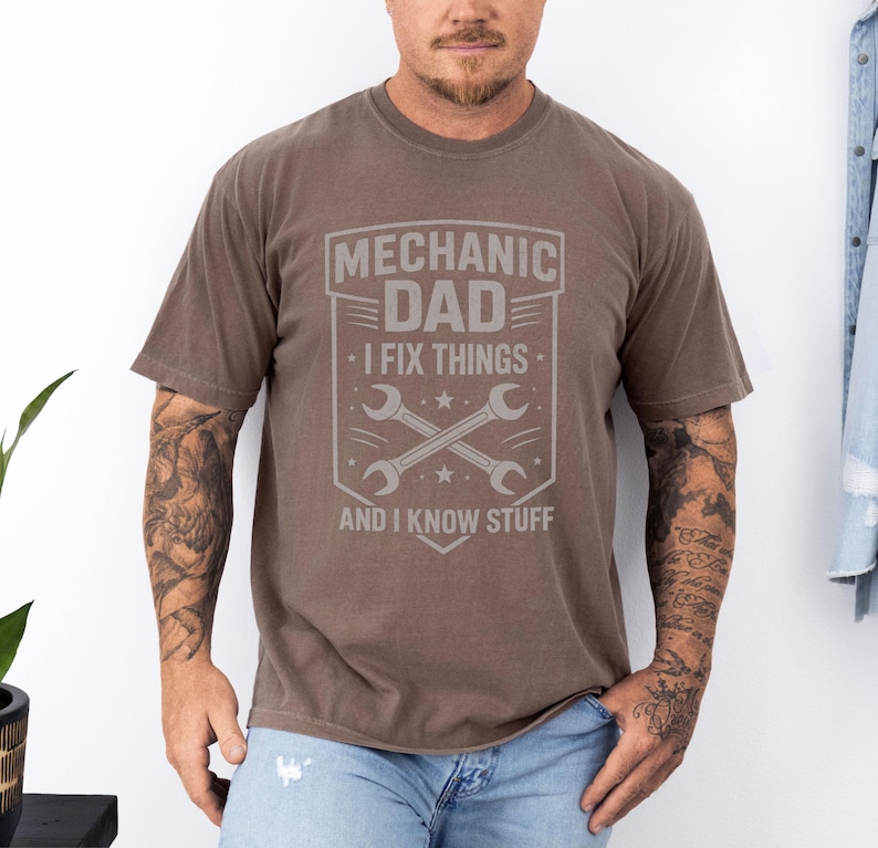 May include: A brown t-shirt with a gray graphic that reads "MECHANIC DAD - I FIX THINGS - AND I KNOW STUFF." The graphic includes crossed wrenches and stars. The shirt has short sleeves and a crew neck.