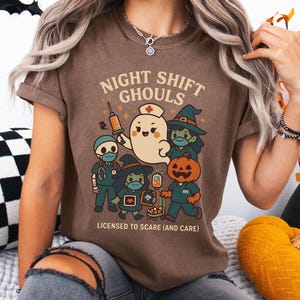 May include: Brown t-shirt with a Halloween-themed graphic. The design features cartoon characters dressed as medical professionals and a ghost, with the text "NIGHT SHIFT GHOULS" and "LICENSED TO SCARE (AND CARE)".
