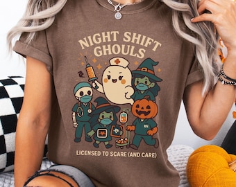 Può includere: T-shirt marrone con grafica a tema Halloween. Il design presenta personaggi dei cartoni animati vestiti da professionisti medici e un fantasma, con la scritta "NIGHT SHIFT GHOULS" e "LICENSED TO SCARE (AND CARE)".