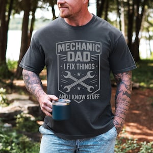 May include: Dark gray t-shirt with the text "MECHANIC DAD I FIX THINGS AND I KNOW STUFF" in a gray graphic. The graphic includes crossed wrenches and stars. The person in the image is holding a blue mug.