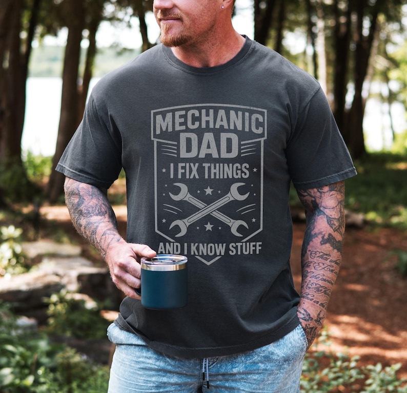 May include: Dark gray t-shirt with the text "MECHANIC DAD I FIX THINGS AND I KNOW STUFF" in a gray graphic. The graphic includes crossed wrenches and stars. The person in the image is holding a blue mug.