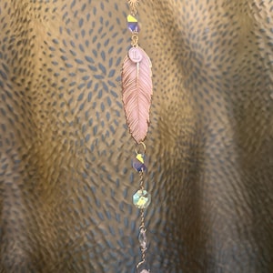 Puede incluir: Un amuleto de pluma rosa con una cadena dorada y cuentas de cristal. La pluma tiene una letra "L" dorada.