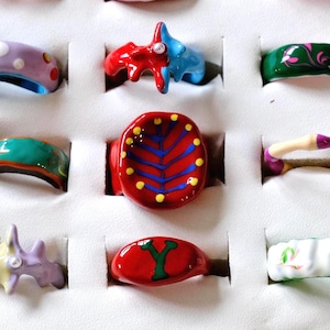 May include: A collection of 10 colorful ceramic rings with various designs, including a tree, a flower, and a geometric pattern. The rings are displayed on a white surface.