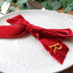 May include: A red velvet ribbon tied in a bow with a gold embroidered letter "R" on a white plate with a decorative pattern.