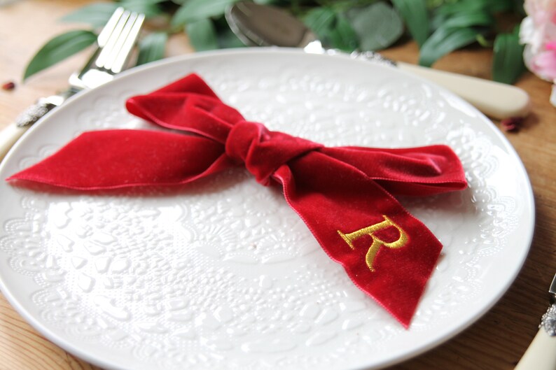 May include: A red velvet ribbon tied in a bow with a gold embroidered letter "R" on a white plate with a decorative pattern.