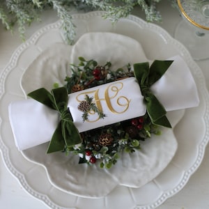 May include: A white napkin with a green velvet bow is tied around a white cylinder with gold embroidery of the letter "H" and a pine sprig. The cylinder is surrounded by a small green wreath with pine cones and red berries. The napkin is on a white plate with a scalloped edge.