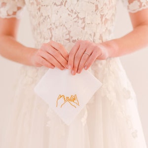 May include: A white handkerchief with a gold embroidered design of two hands making a pinky promise.