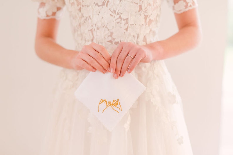 May include: A white handkerchief with a gold embroidered design of two hands making a pinky promise.