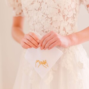 May include: A white handkerchief with a gold embroidered design of two hands making a pinky promise.