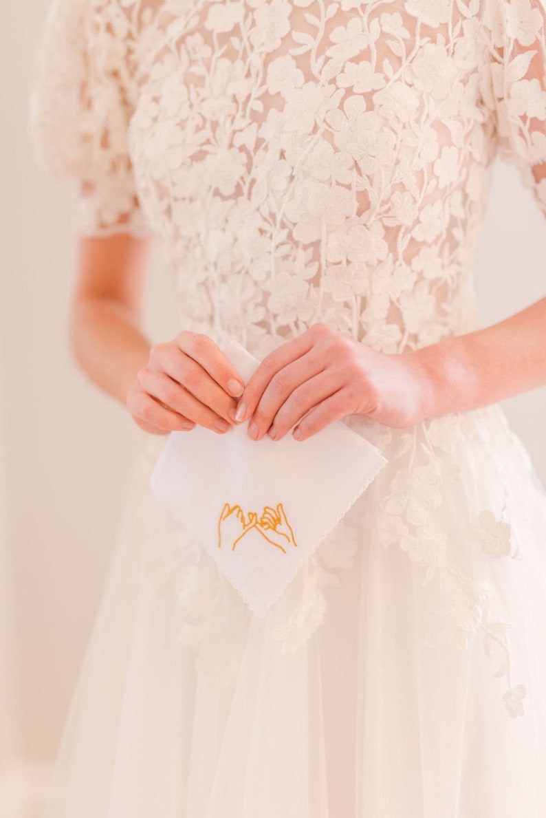 May include: A white handkerchief with a gold embroidered design of two hands making a pinky promise.