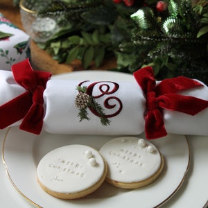 May include: A white Christmas cracker with a red velvet bow and a monogrammed "C" embroidered on the front. Two white cookies with the text "Merry Christmas" and "Happy Christmas" are on a white plate.