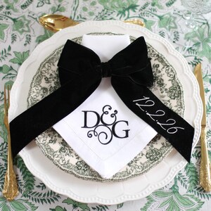 Puede incluir: Una servilleta de lino blanco con una cinta de terciopelo negro atada en un lazo. La servilleta está bordada con las iniciales "D&G" y la fecha "12.2.26".