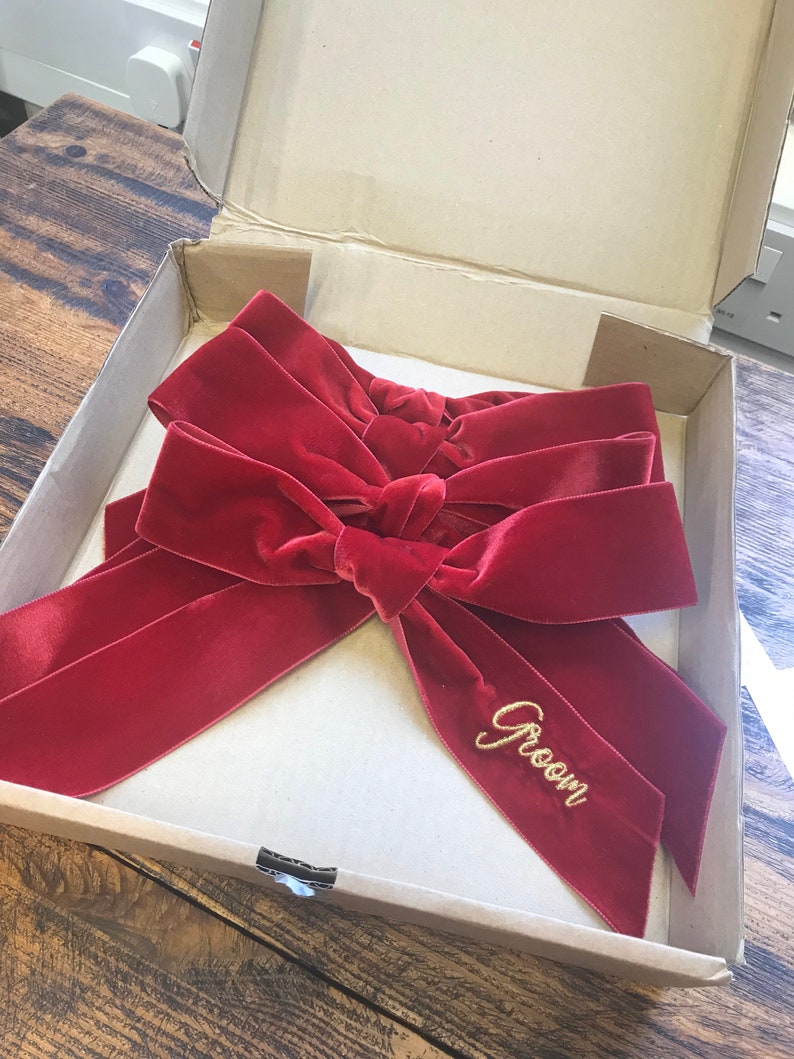 Op de afbeelding: Een rode fluwelen strik met het woord "Groom" geborduurd in gouden draad. De strik zit in een kartonnen doos.