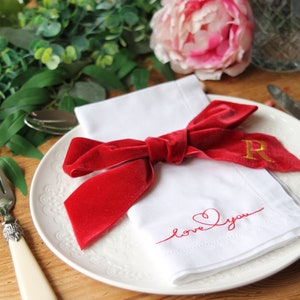 May include: A white napkin with a red velvet ribbon tied in a bow and the words "love you" embroidered in red thread. The napkin is on a white plate with a gold letter "R" embroidered on the ribbon.