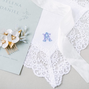 May include: A white lace-trimmed handkerchief featuring a blue and purple embroidered monogram, a floral hair comb with white petals and gold leaves, and a light green card with the printed text Selena and Daniel.