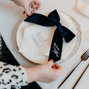 Op de afbeelding: Een witte servet met een gebouwillustratie en de naam "Lily" in cursief op een donkerblauw lint dat in een strik is gebonden. De servet ligt op een wit bord met een geweven placemat.