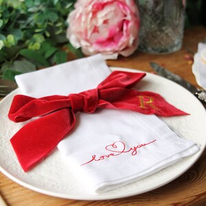 May include: A white napkin with a red velvet bow and the words "love you" embroidered in red thread. The napkin is on a white plate with a gold initial "R" embroidered on the bow.