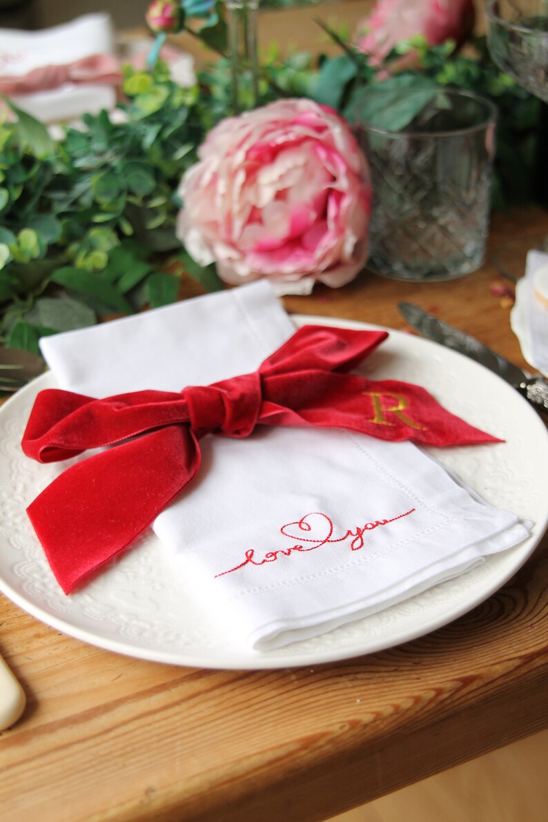 May include: A white napkin with a red velvet bow and the words "love you" embroidered in red thread. The napkin is on a white plate with a gold initial "R" embroidered on the bow.
