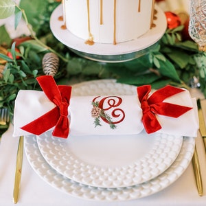 May include: A white napkin with a red velvet bow and the letter "E" embroidered on it. The napkin is placed on a white plate with a textured design. The plate is on a white tablecloth with gold silverware.