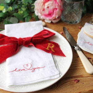 May include: A white napkin with red embroidery that says "love you" with a heart. A red velvet bow is tied around the napkin with a gold embroidered "R" on the bow. The napkin is on a white plate with a decorative edge. A white knife with a cream handle is on the table next to the plate.