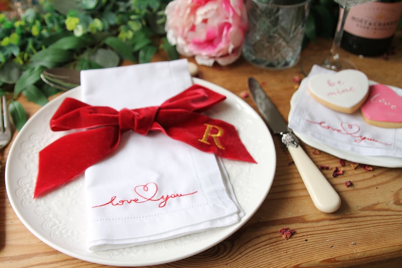 May include: A white napkin with red embroidery that says "love you" with a heart. A red velvet bow is tied around the napkin with a gold embroidered "R" on the bow. The napkin is on a white plate with a decorative edge. A white knife with a cream handle is on the table next to the plate.