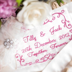 Puede incluir: Jarretera de satén blanco con encaje rosa y un mensaje bordado en rosa que dice "Tilly & Jack 26 de diciembre de 2012 Juntos para siempre".