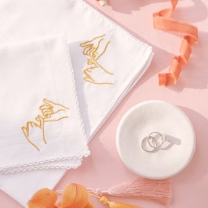 May include: Two white handkerchiefs with gold embroidery of hands holding each other. The handkerchiefs are on a pink surface with a small white dish containing two silver rings, a peach-coloured ribbon, and a peach-coloured orchid with orange tassels.