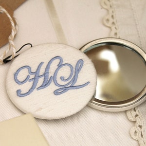 May include: A white fabric compact mirror with a blue embroidered monogram "HL" on the front. The mirror is open and the back is silver.