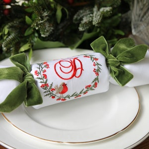 May include: A white fabric Christmas cracker with a green velvet bow. The cracker is decorated with an embroidered wreath of holly and berries, a robin, and the letter "D".